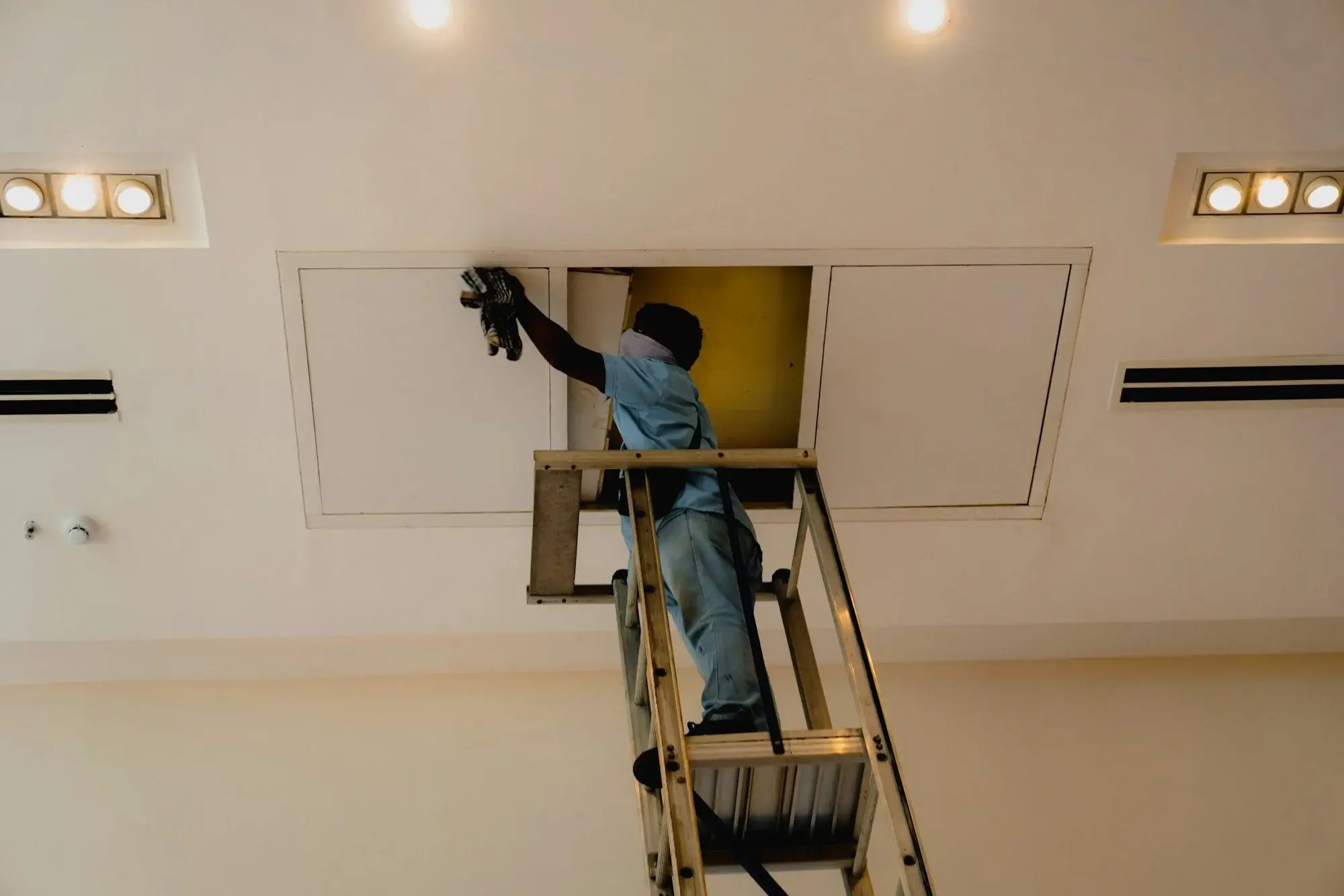 Técnico de manutenção predial em uma escada abrindo o alçapão do forro de um prédio comercial